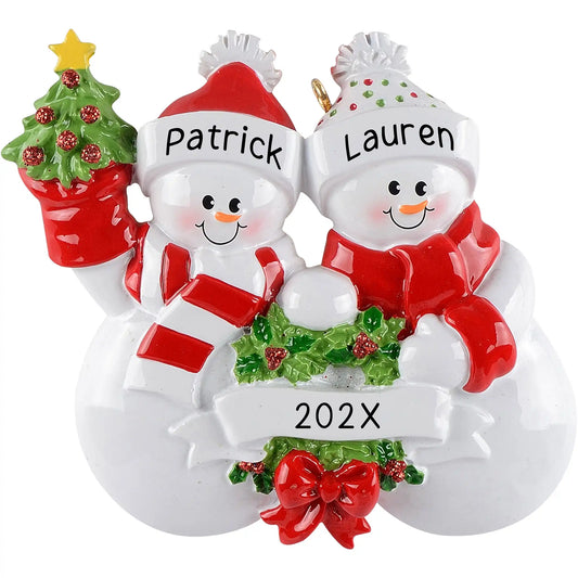 Personalized Christmas ornament featuring a snowman couple in red and white winter hats, holding a miniature Christmas tree and garland, with a blank white banner for custom name engraving.