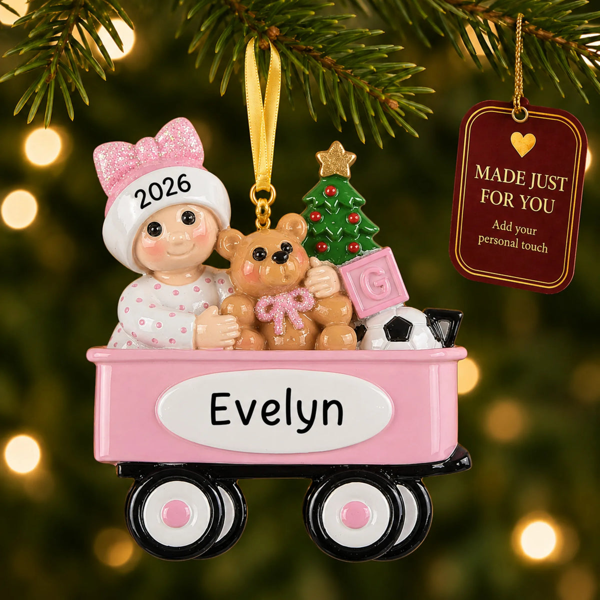 Pink wagon baby girl christmas ornament hanging on a lit Christmas tree with a Made Just For You gift tag