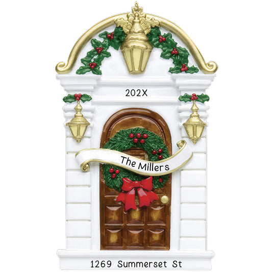 A personalized holiday ornament featuring a brown house door with a wreath, holly leaves, and a bow. The front door has 'The Millers' written on it, and the year 'Year' at the top.