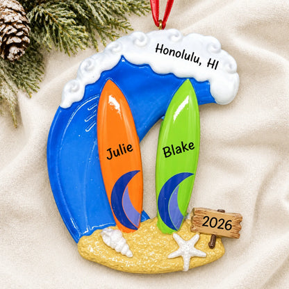 Personalized big wave surfing couple christmas ornament laid on a soft white knit blanket