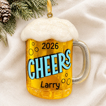 Personalized beer mug Christmas ornament reading Cheers, Larry, and 2026 on a white knit blanket with pine