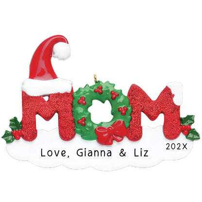 A personalized Christmas ornament with the word 'Mom' in red and green, surrounded by holly and berries, and a Santa hat. It has been customized with the names 'Gianna & Liz' and the year '202X'.