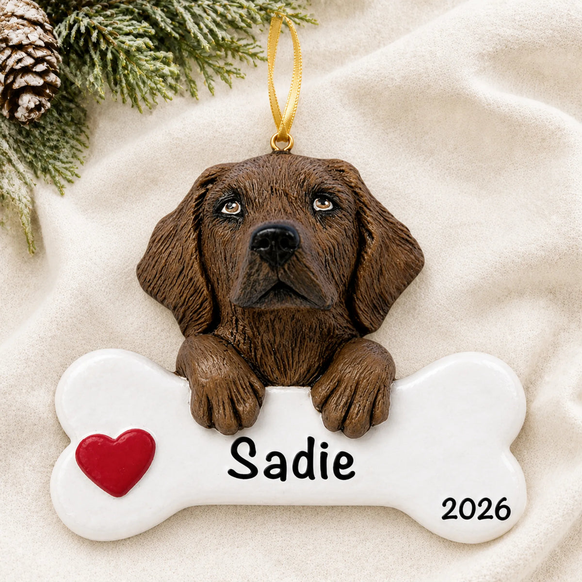 Personalized chocolate Lab dog bone Christmas ornament resting on a soft white blanket with pine and pinecones
