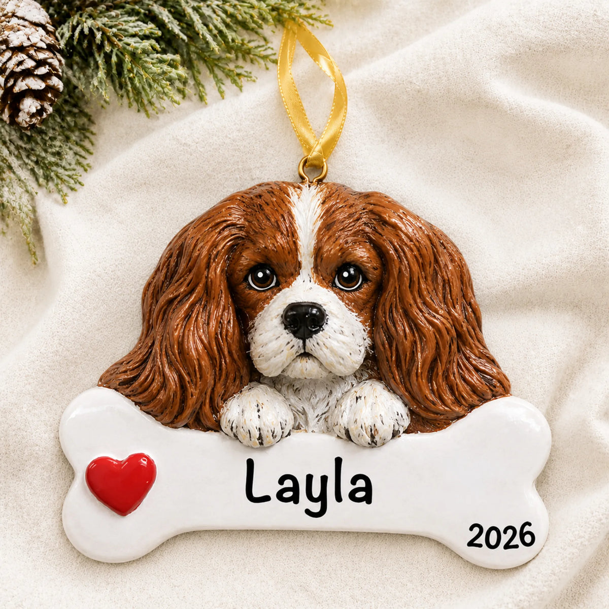 Personalized King Charles Cavalier dog bone Christmas ornament resting on a soft white blanket with pine and pinecones