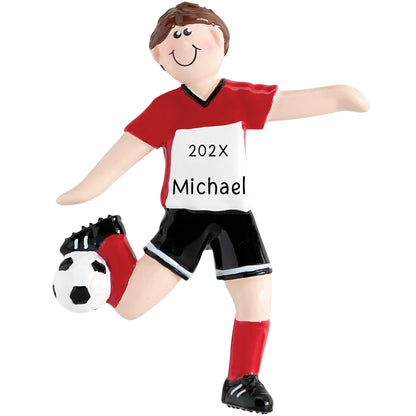A personalized holiday ornament depicting a boy in a red and white soccer uniform with a ball and shin guards. The ornament has a smiling face and the text '202x Michael' on the front.