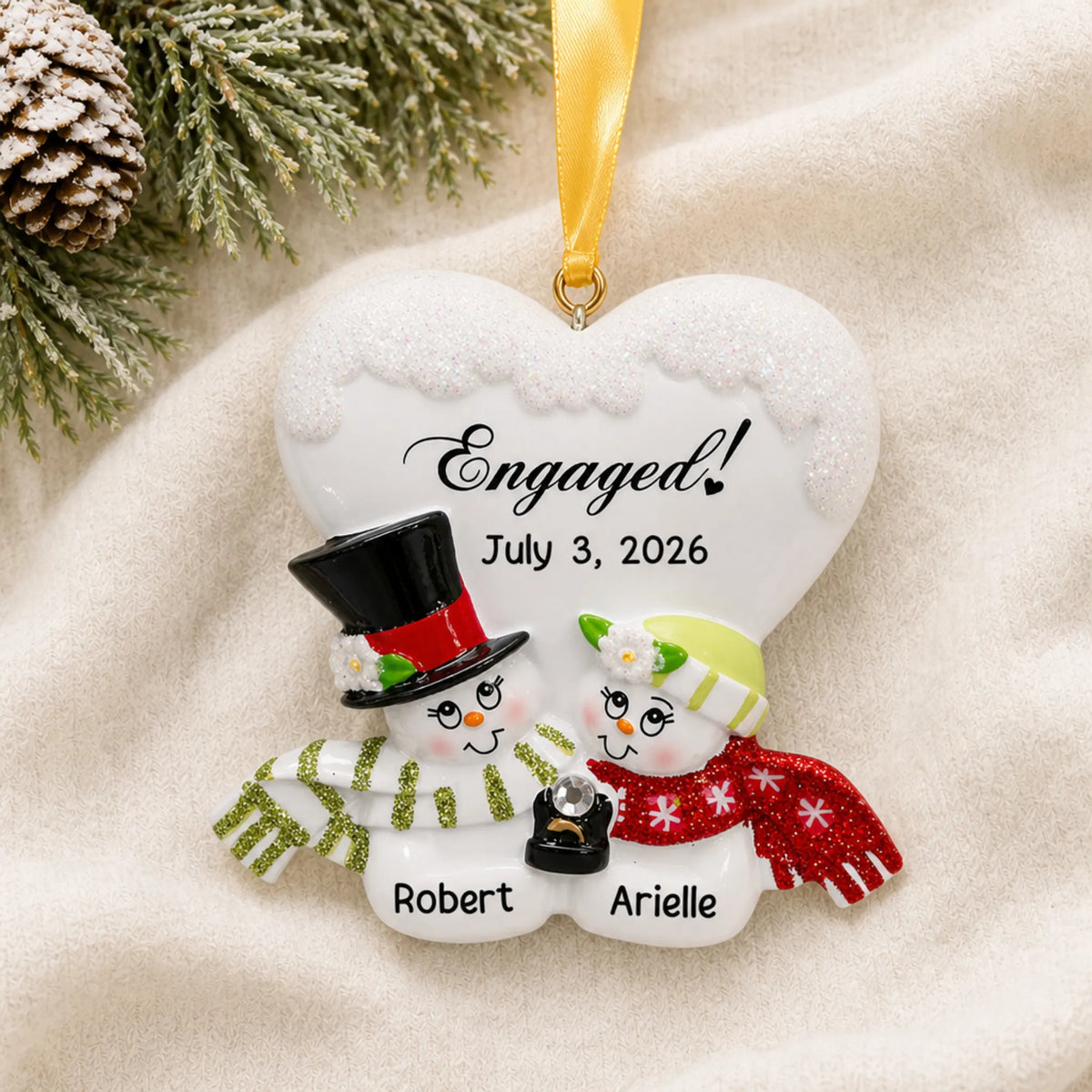 Personalized engaged snow couple Christmas ornament on a cream knit blanket beside pine and pinecones