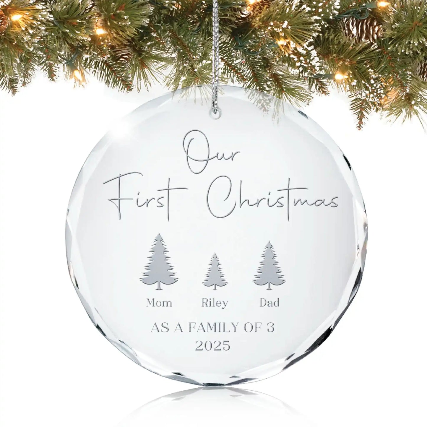 Personalized glass ornament engraved with “Our First Christmas as a Family of 3,” names under three trees, and year 2025, hanging on a Christmas tree.