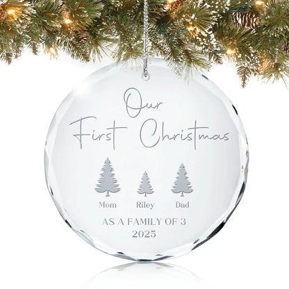 Personalized glass ornament engraved with “Our First Christmas as a Family of 3,” names under three trees, and year 2025, hanging on a Christmas tree.