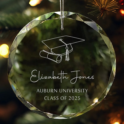 Personalized glass graduation ornament engraved with a cap, diploma, and “Class of 2025” hanging on a Christmas tree.