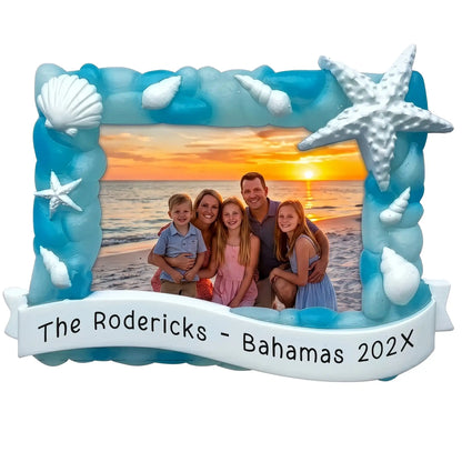 Personalized beach photo frame Christmas ornament with a sea glass blue shell-and-starfish frame holding a family beach photo, and a banner engraved with a custom name, destination, and year.