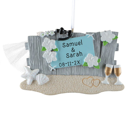 Personalized beach wedding Christmas ornament, a driftwood sign on the sand with a starfish, champagne flutes, and a bridal veil, engraved Samuel & Sarah with their wedding date
