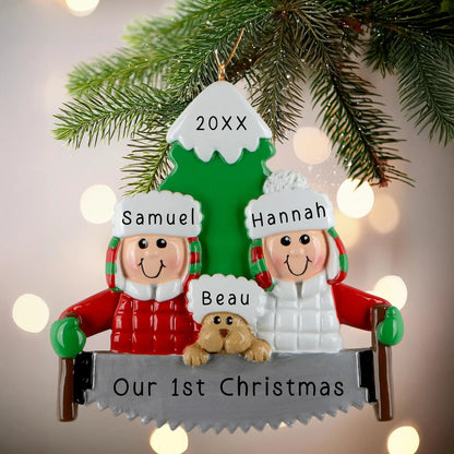 A personalized resin ornament featuring a couple in winter clothing with a dog, standing in front of a Christmas tree. The ornament includes customizable text for names and a year. The ornament is attached to a Christmas tree with a festive look.