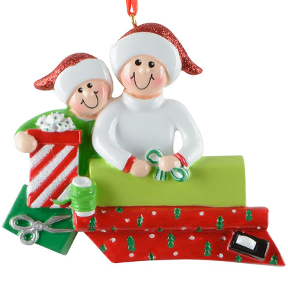 A personalized Christmas ornament of two people in Santa hats wrapping a gift, featuring a green box for custom engraved names.