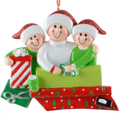 A personalized Christmas ornament of three people in Santa hats wrapping holiday gifts, featuring a green box for custom engraved names.