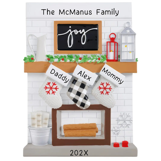 A table top Christmas ornament featuring a fireplace mantle with three customizable stockings. The ornament has a 'The McManus Family' text above the fireplace, and each stocking can be personalized with names and a year below.