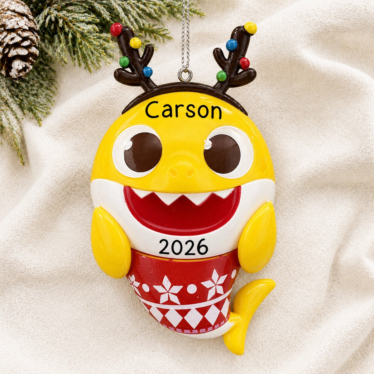 Personalized Baby Shark Christmas ornament reading Carson 2026, resting on a white knit blanket with pine and a pinecone