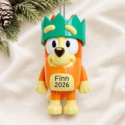 Personalized Bingo with crown Christmas ornament reading Finn 2026, resting on a white knit blanket with pine and a pinecone
