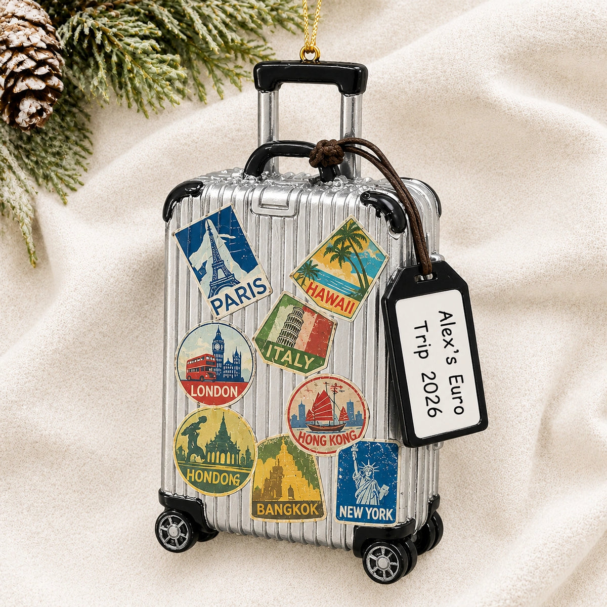 Personalized travel suitcase Christmas ornament reading Alex's Euro Trip 2026 on a white knit blanket with pine