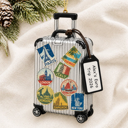 Personalized travel suitcase Christmas ornament reading Alex's Euro Trip 2026 on a white knit blanket with pine