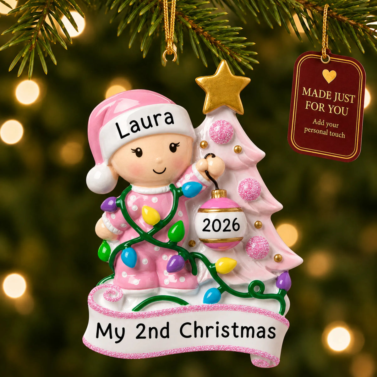 Pink baby's 2nd christmas ornament hanging on a lit Christmas tree with a Made Just For You gift tag