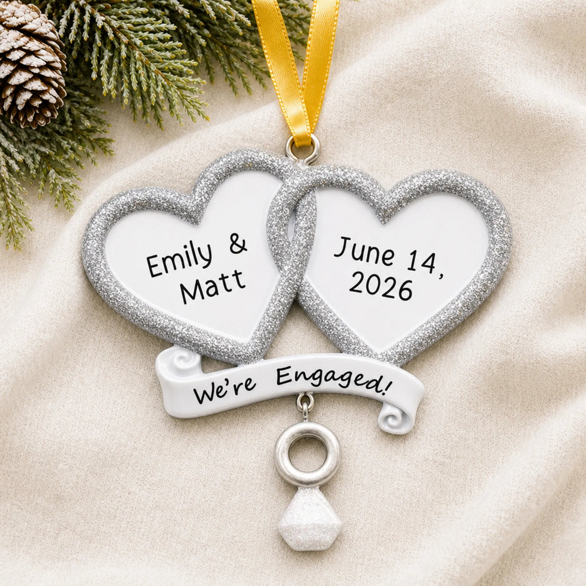 Personalized We're Engaged double heart Christmas ornament on a cream knit blanket beside pine and pinecones