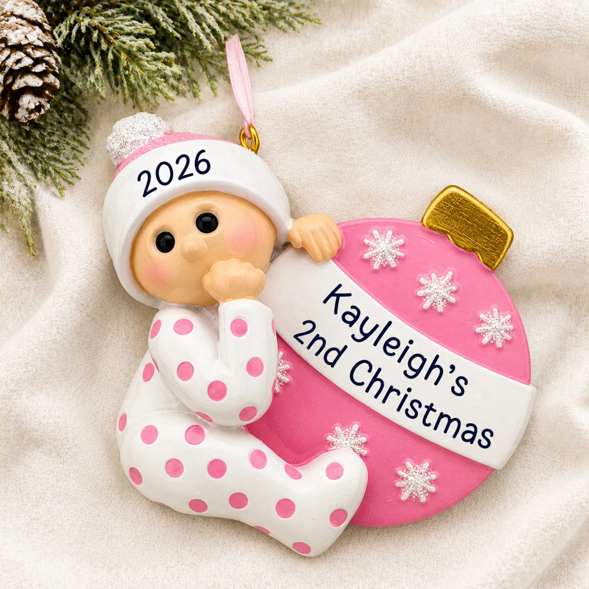 Personalized pink baby's 2nd christmas ornament laid on a soft white knit blanket